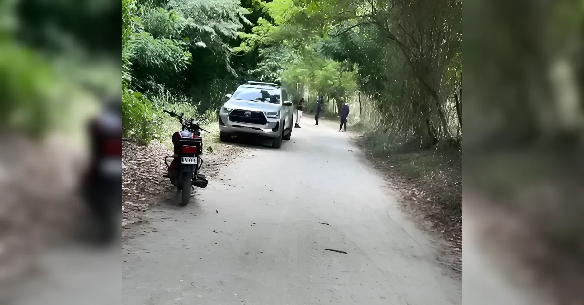 Dirt road in a wooded area with a white SUV and a motorcycle parked on the left; several people stand ahead.