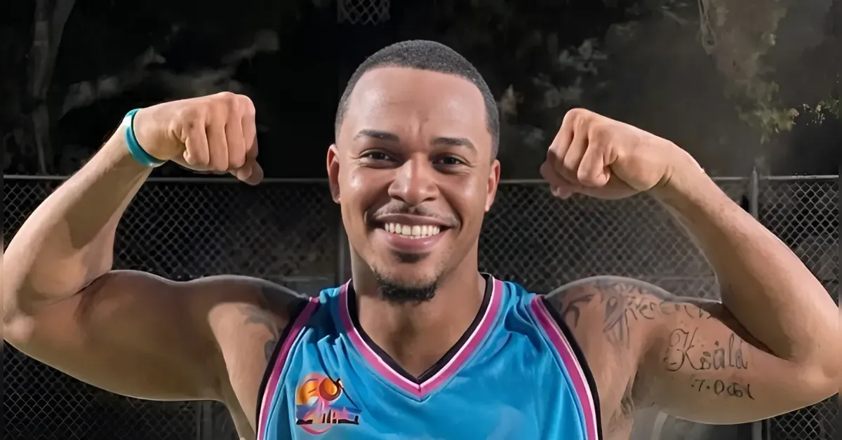 Smiling man in a blue basketball jersey flexing his biceps outdoors on a fenced court at night, tattoos visible on his arms.