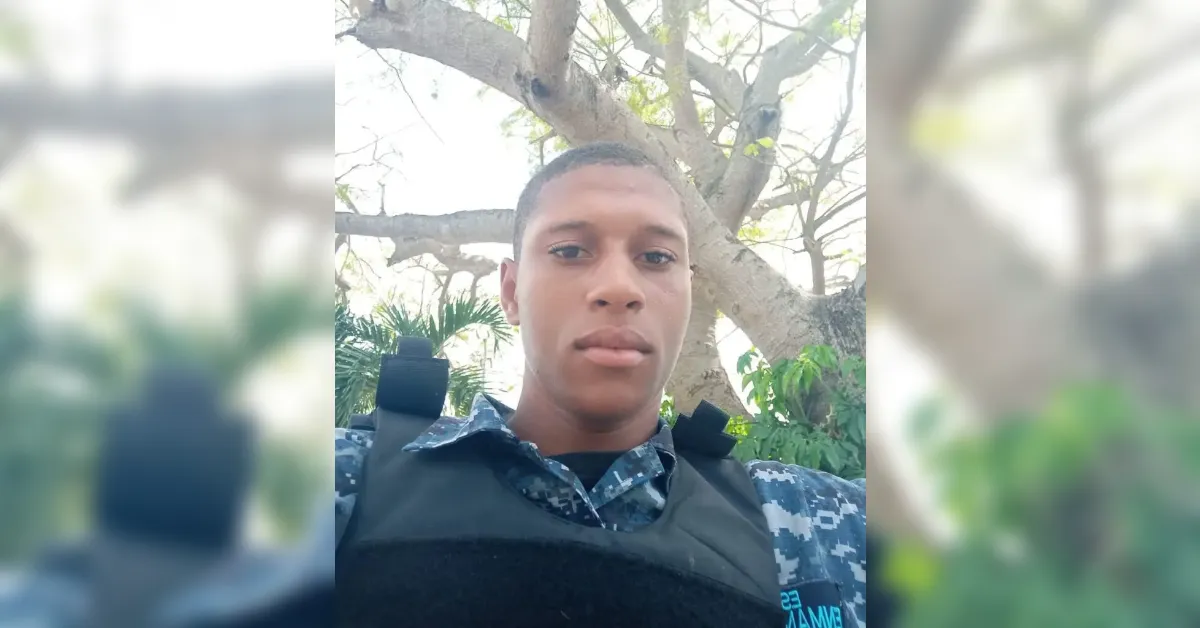 Young man in camouflage uniform and tactical vest taking a selfie outdoors with a large tree behind him.