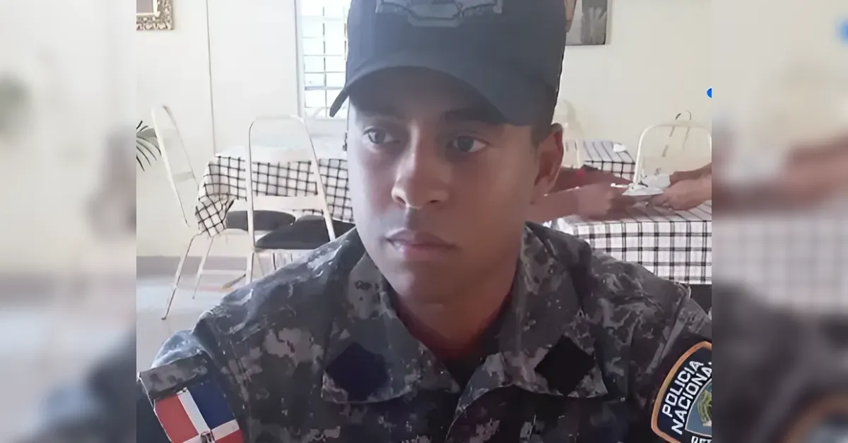 Close-up of a young man in camouflage military uniform and cap, indoor setting with checkered tablecloths in the background; visible flag patch on the right sleeve and a Police Nacional badge on the left sleeve.