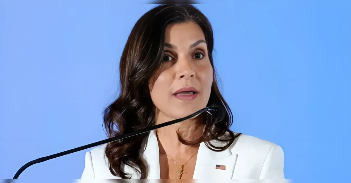 Woman in a white blazer speaking at a podium with a microphone, against a light blue background, wearing a small American flag pin and a gold necklace.