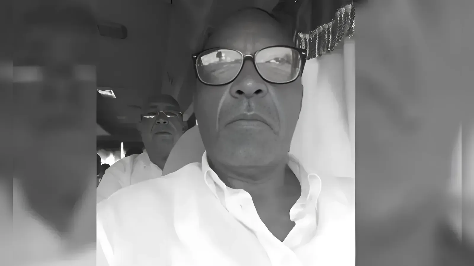 Close-up of an older man wearing glasses and a white shirt, looking at the camera with a serious expression; a second man is faintly visible in the background.