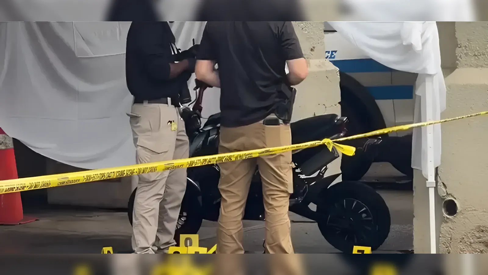 Investigators examine a black motorcycle behind yellow caution tape at a taped-off crime scene.
