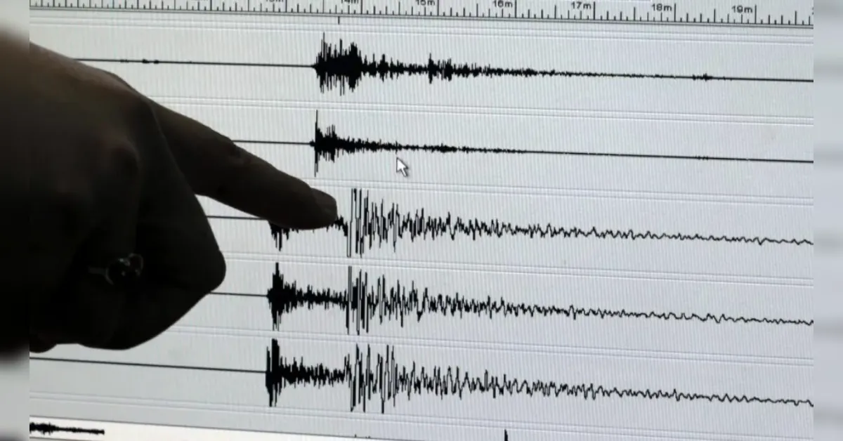 Person's finger points at a waveform on a computer monitor, showing multiple audio/seismic tracks with a spike in the center edge of the screen.