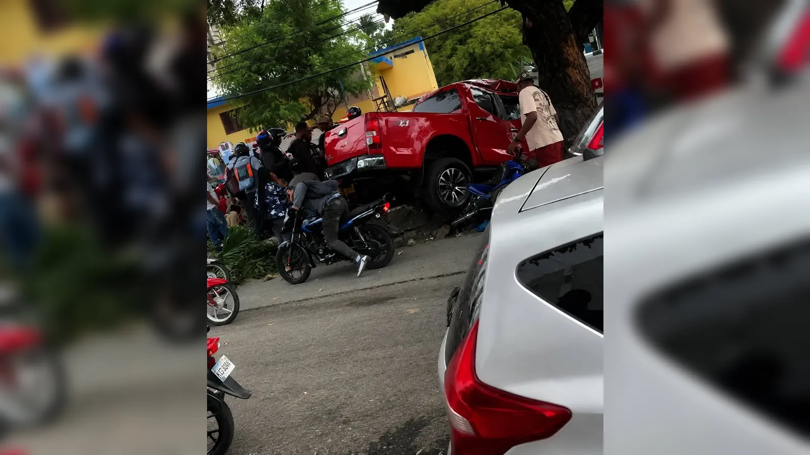 Red pickup truck on a curb with its rear raised, surrounded by bystanders and motorcycles on a city street area.