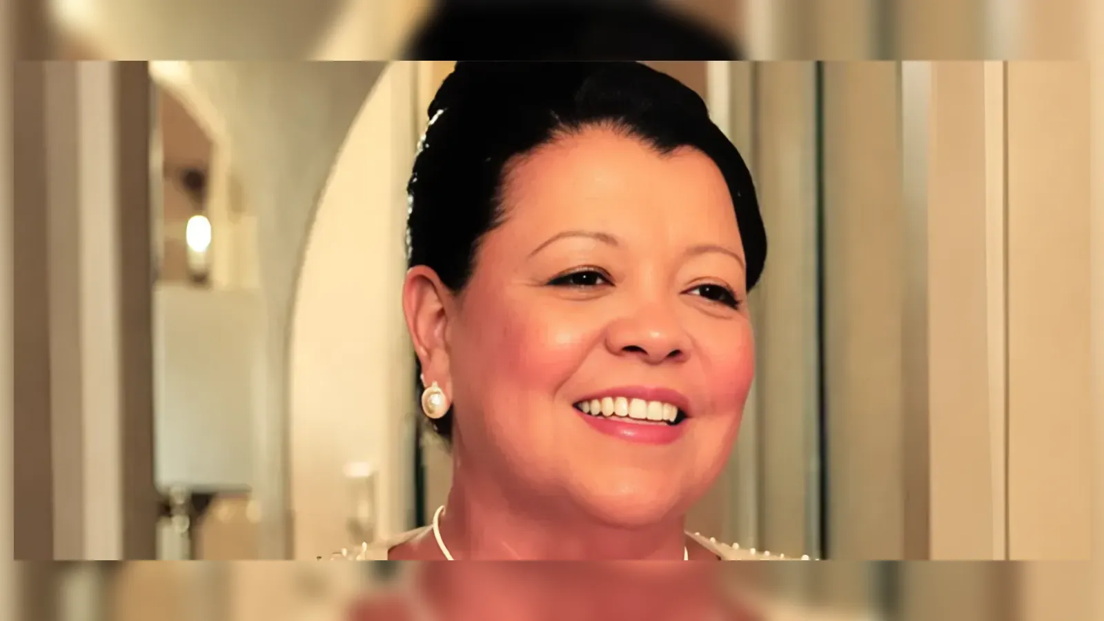 Smiling woman with dark hair in an updo, wearing pearl earrings, indoors.