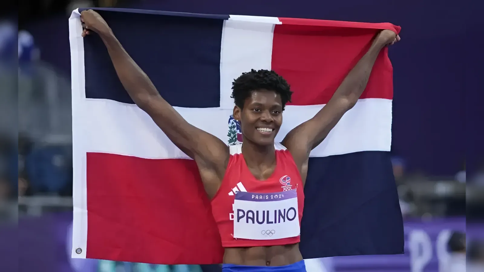 Olympic athlete Paulino raises a Dominican Republic flag in celebration, wearing a red top and blue shorts on the podium.