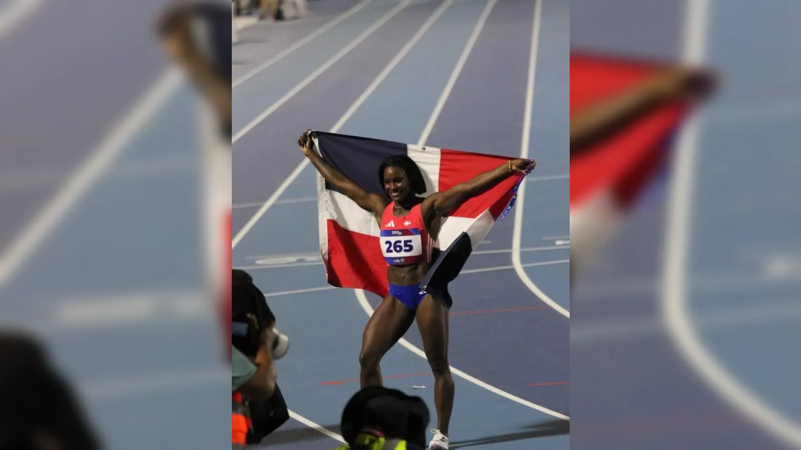 Athlete crosses the track finish line with arms raised, holding a blue-white-red flag aloft behind them in celebration.