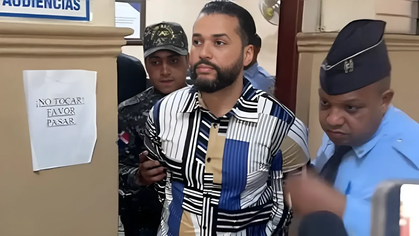 Bearded man in a geometric black‑white‑blue shirt is escorted by security guards through a doorway.