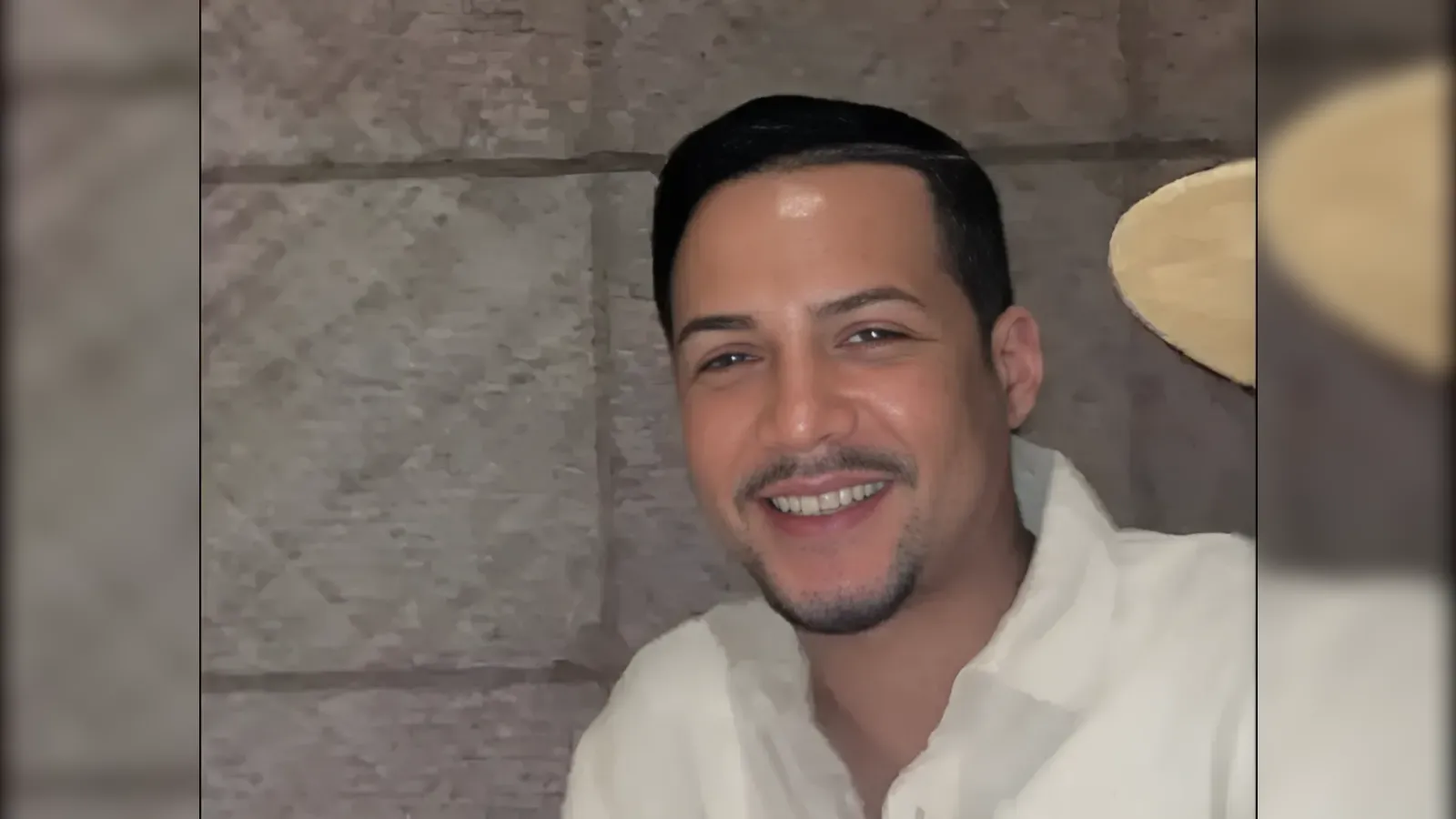 Smiling man with short dark hair and light stubble wearing a white shirt, posed against a textured stone wall background.