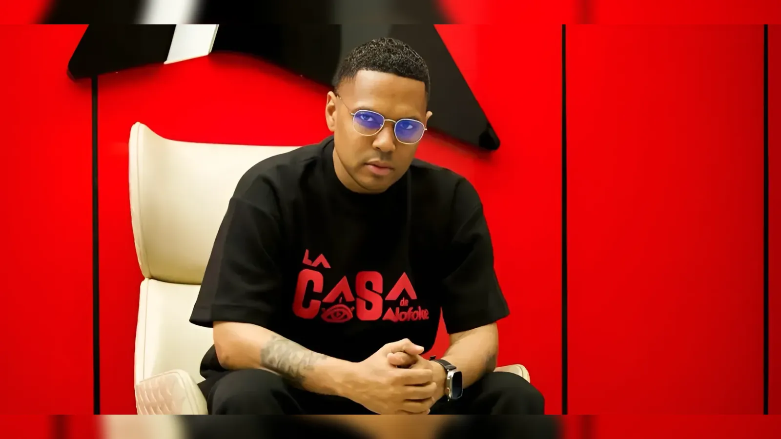 Man seated in a white chair against a bright red wall, wearing a black shirt and blue-tinted sunglasses, with his hands clasped.