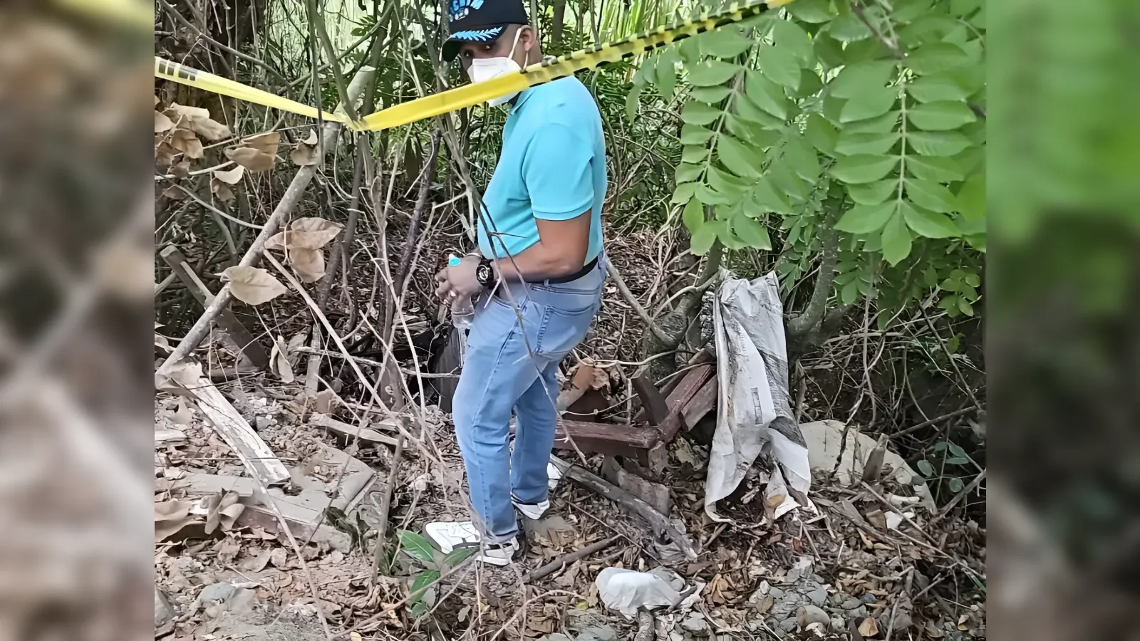 Man in a teal shirt and jeans wearing a cap and mask stands among debris and overgrown bushes, with yellow caution tape crossing the scene.