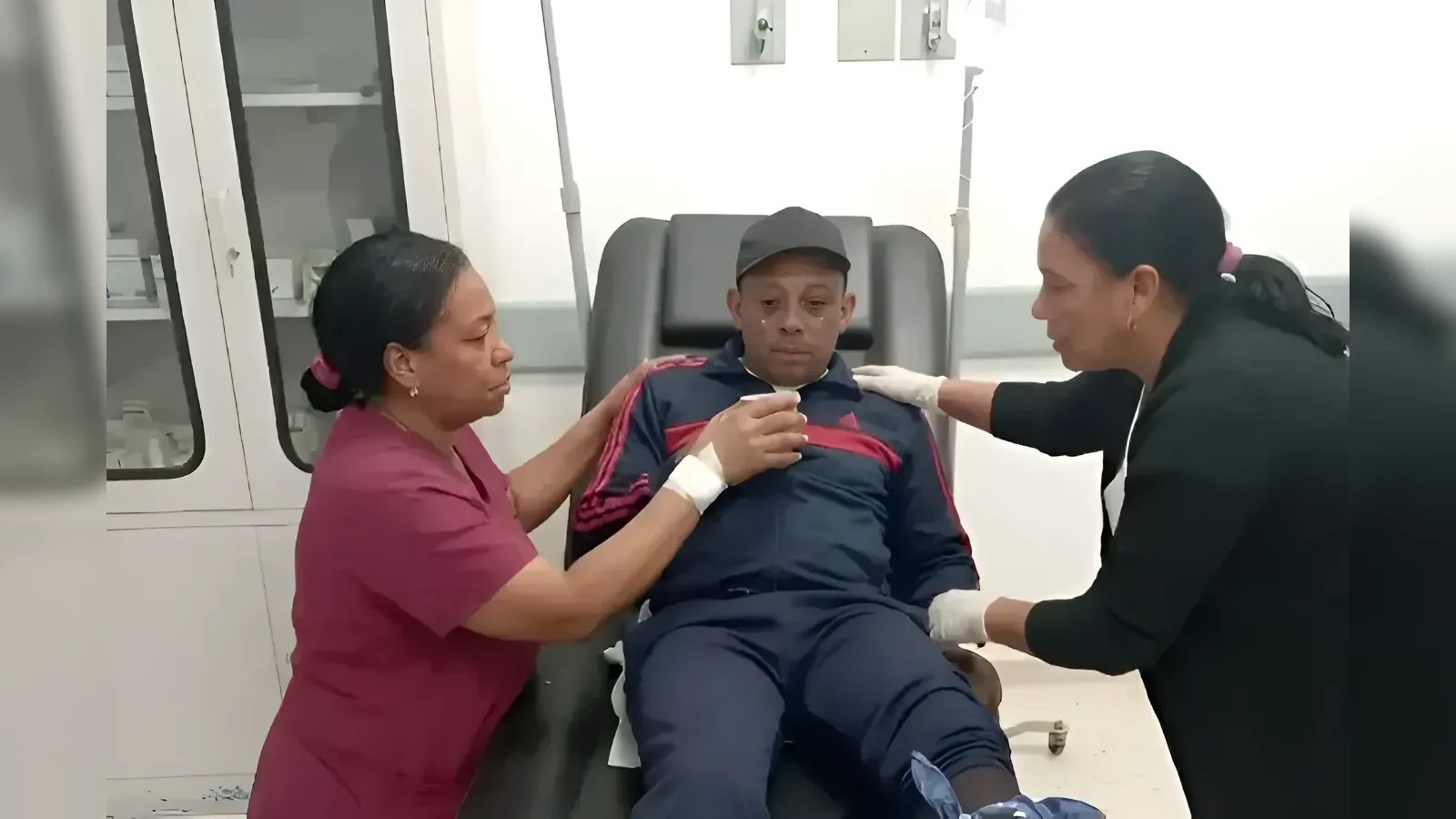 Man reclining on a medical chair being assisted by two caregivers in a clinic; one holds his hand while the other rests a hand on his shoulder.
