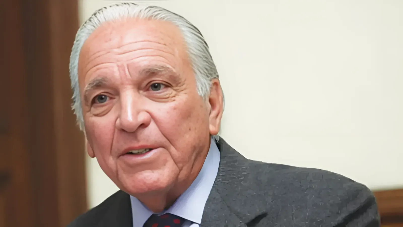 Close-up of an older man with white hair wearing a dark suit and light blue shirt, looking slightly to the side in an indoor setting