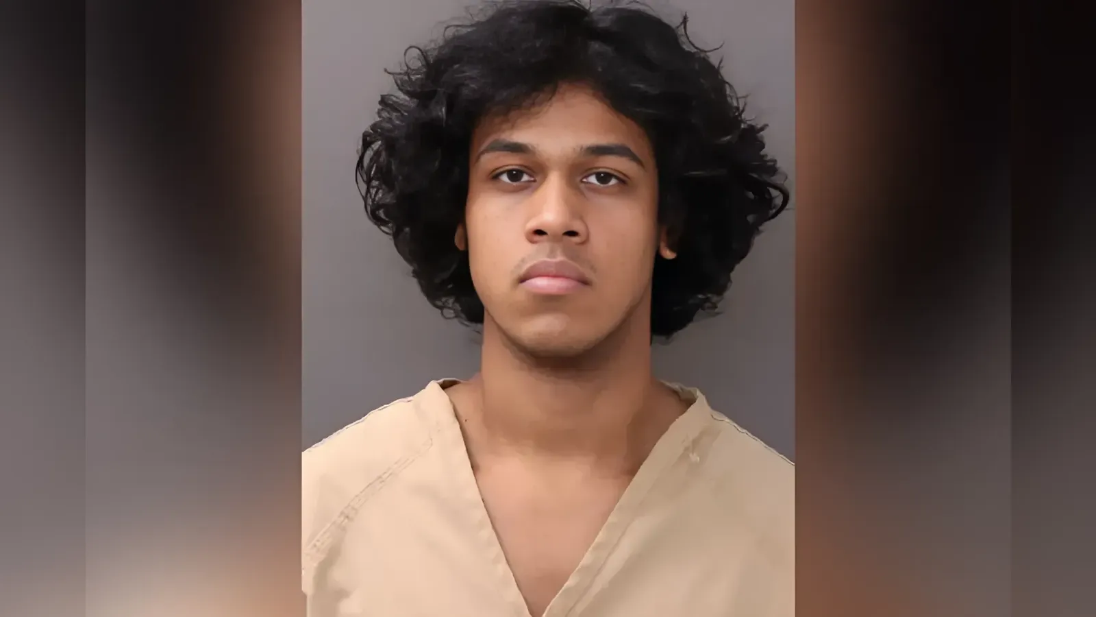Young man with curly black hair in a beige prison uniform against a gray backdrop in a mugshot-style photo.