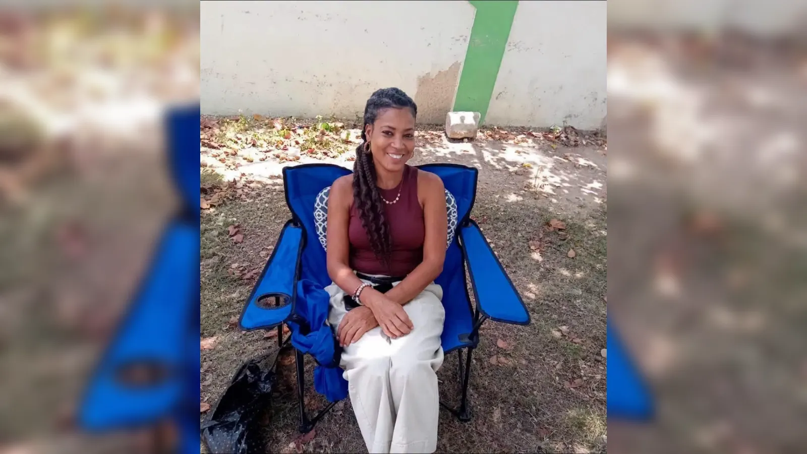 Woman seated in a blue folding chair outdoors, smiling, wearing a maroon sleeveless top and white pants, with braided hair.