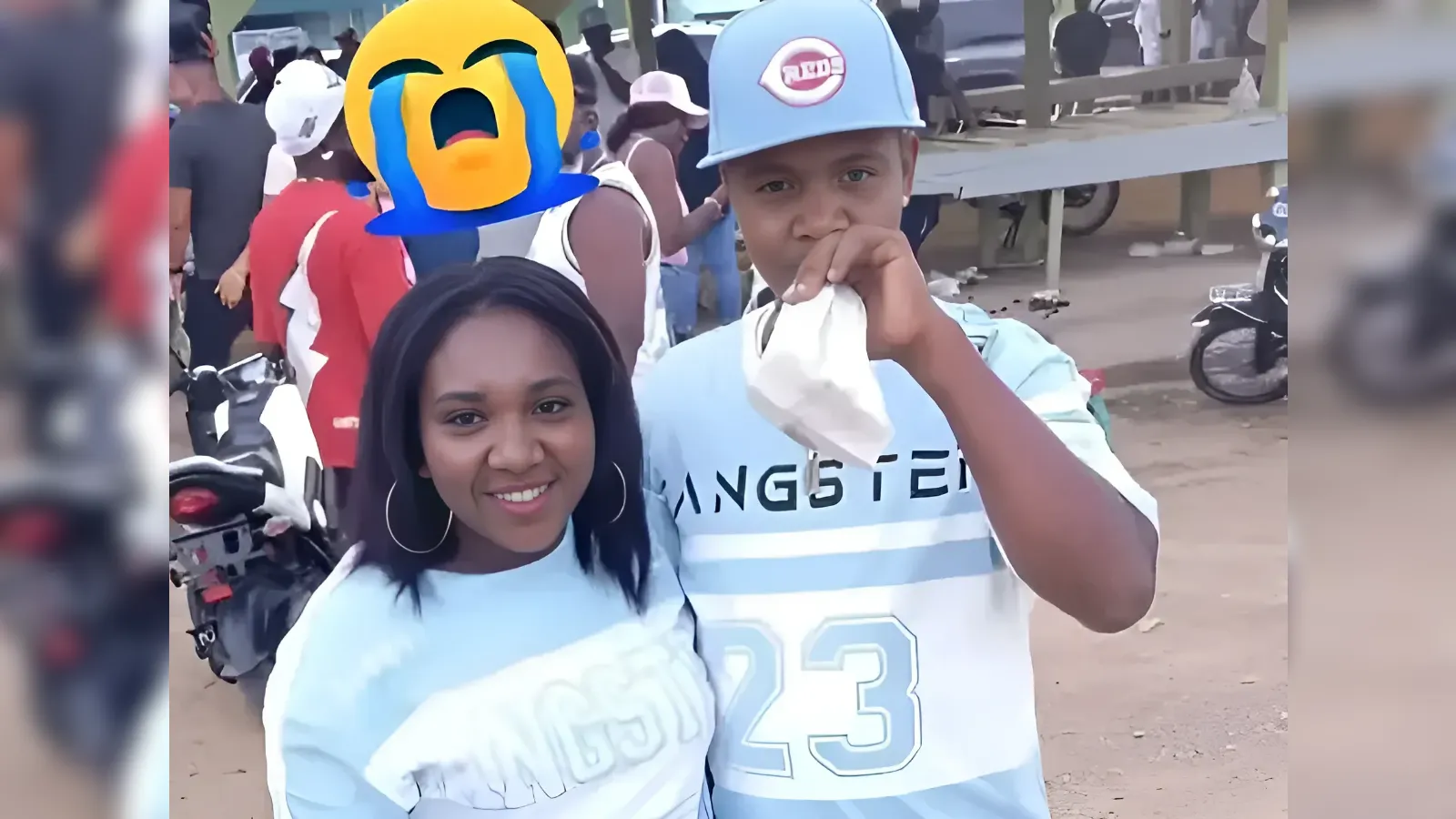 Two friends posing outdoors at a crowded event; man in a light-blue jersey #23 wipes his mouth with a tissue as a yellow crying emoji appears in the background.