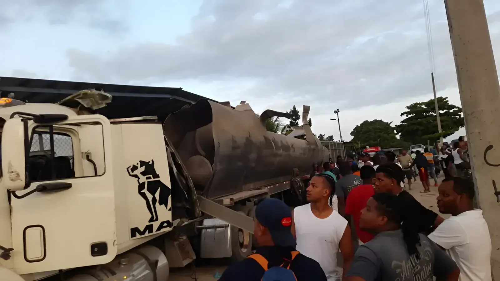 White Mack tanker truck with a crushed cylindrical tank, crowd of onlookers gathered beside it at what looks like an accident scene