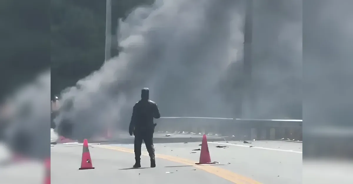 Person in dark protective gear stands on a road between orange cones as thick smoke fills the scene, with debris scattered nearby.