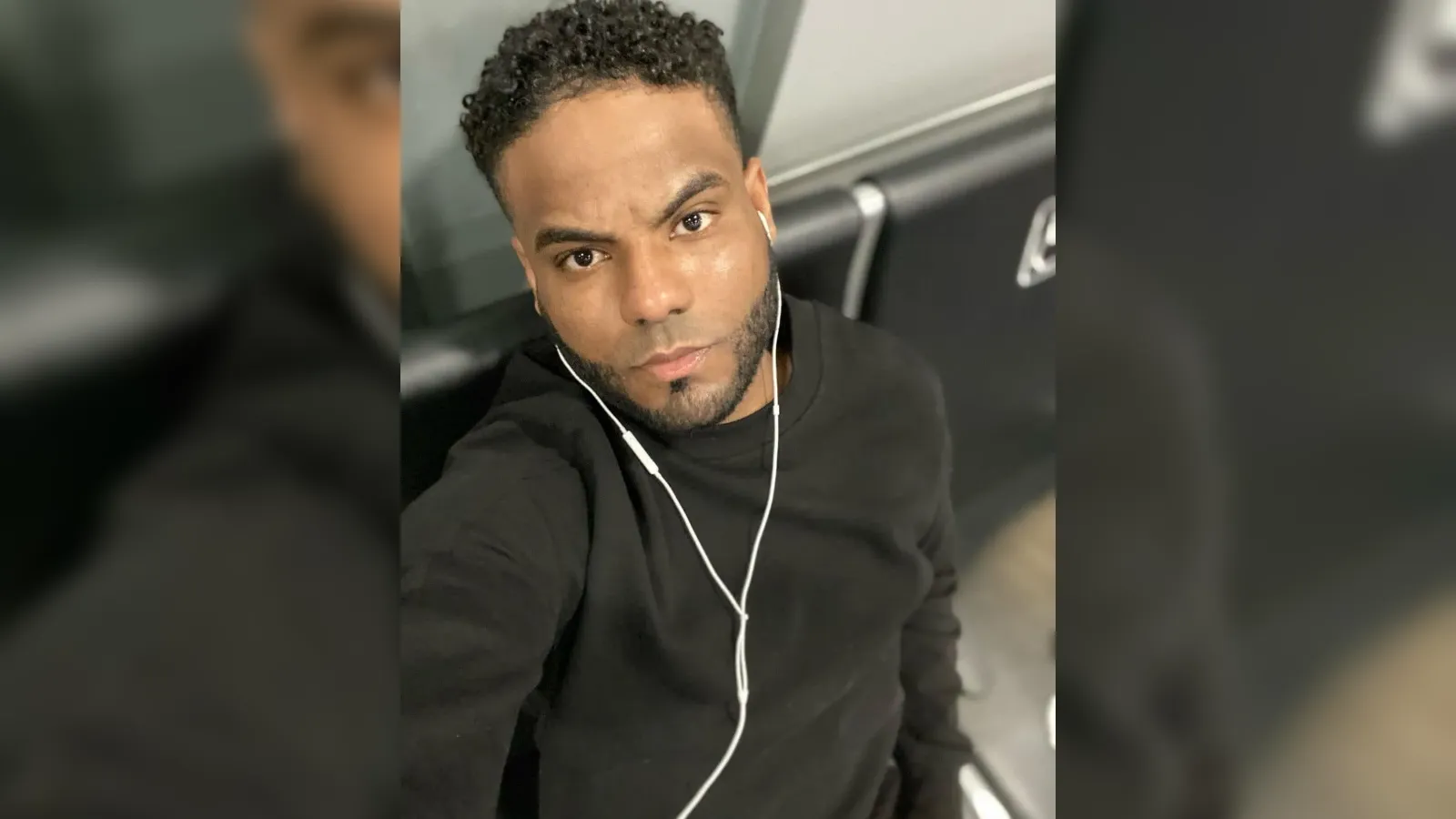 Self-portrait of a man with short curly hair, wearing a black long-sleeve shirt and white earbuds, looking at the camera to take a selfie.