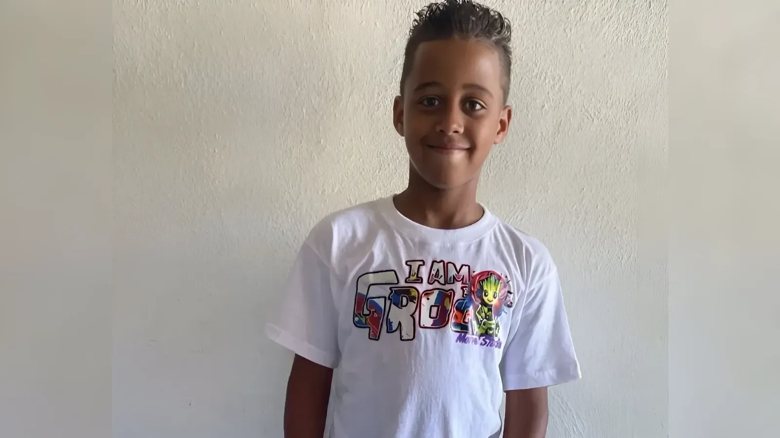 Smiling boy in a white T-shirt with an 'I AM GROOT' graphic, standing against a light textured wall.
