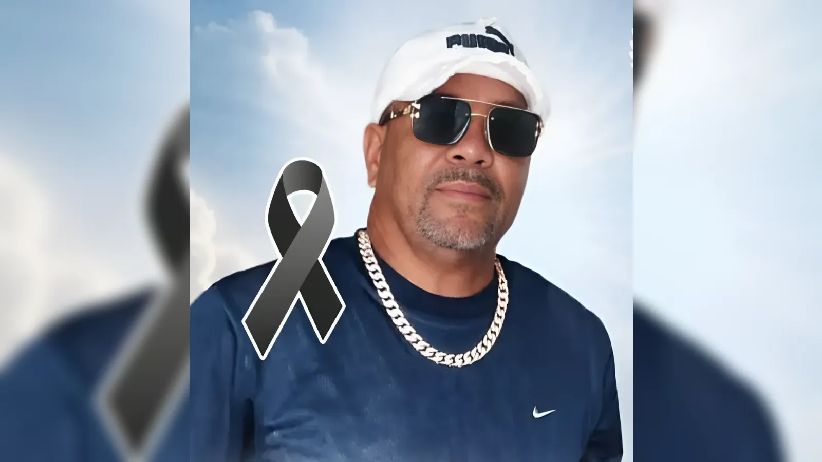 Portrait of a man in sunglasses and a white head wrap, blue shirt, and silver chain; a gray mourning ribbon indicates a memorial tribute.