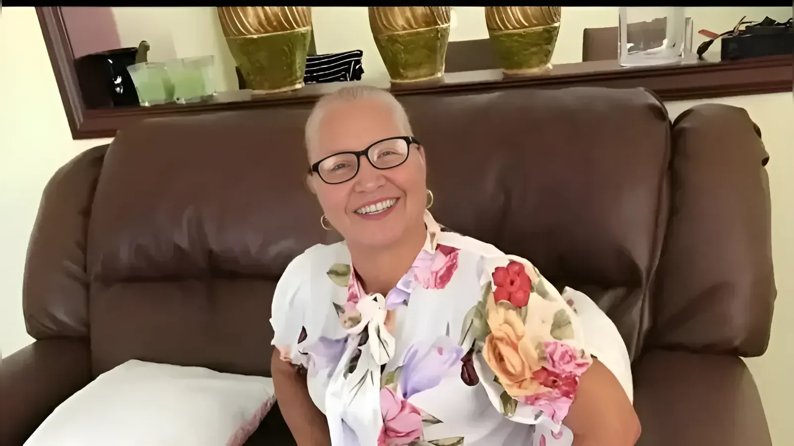 Smiling older woman with short white hair and black glasses sits on a brown leather couch, wearing a white floral blouse.