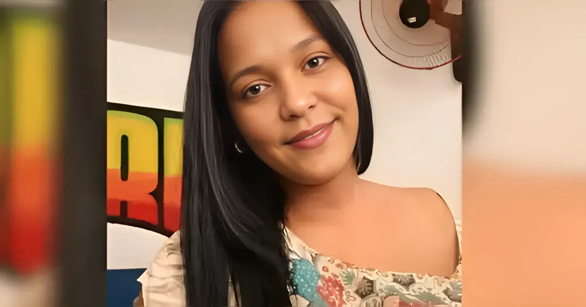 Smiling woman with long black hair in a floral off-shoulder top, indoors in front of a colorful backdrop on a video frame.