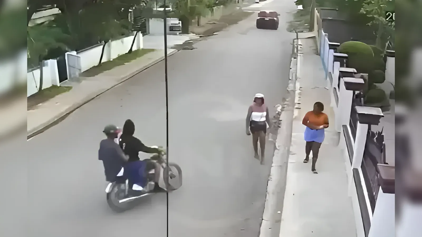Two-person motorcycle riding down a quiet street, with pedestrians on the sidewalk ahead near a fenced yard edge.
