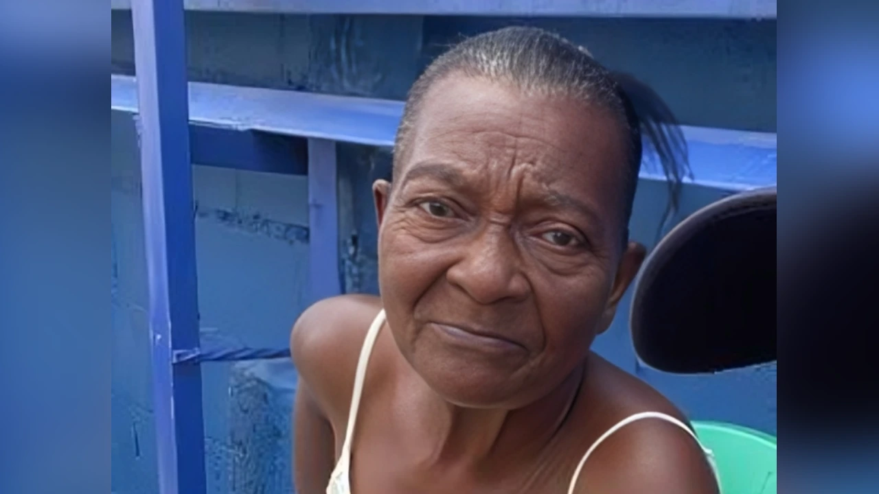 Close-up of a smiling older woman with slicked-back hair wearing a white tank top, standing beside a blue railing.