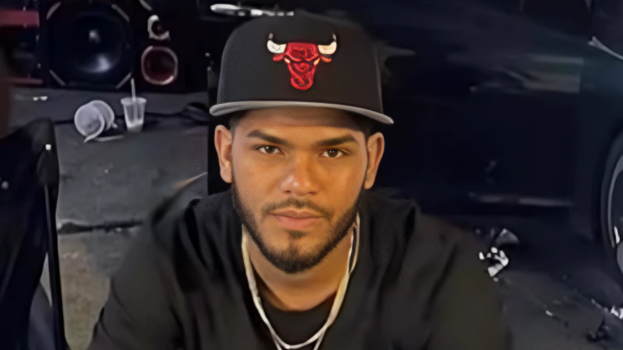 Portrait of a man wearing a black cap with a red Chicago Bulls logo, looking at the camera in a dim indoor setting near a car.