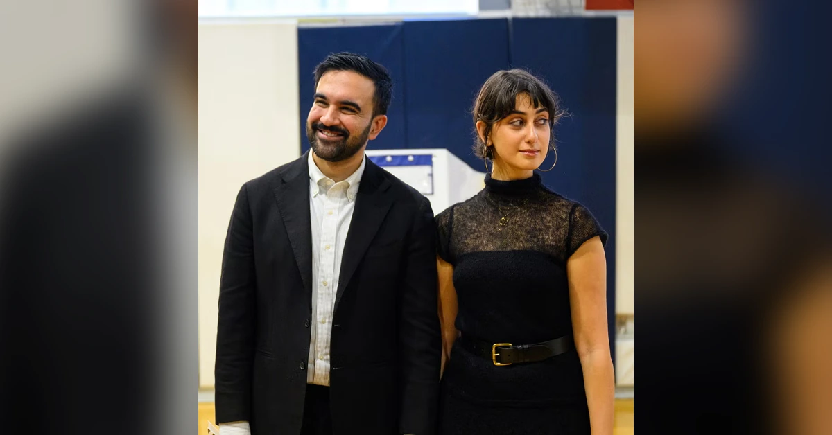 Man in a black suit and white shirt smiles beside a woman in a black lace dress with a belt, standing indoors at an event.