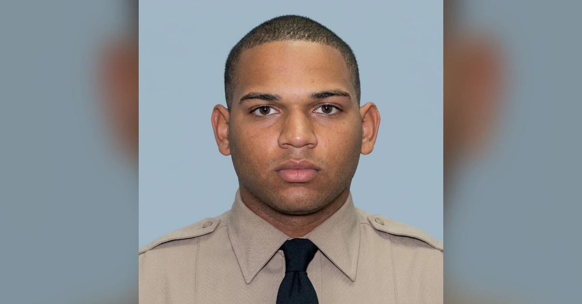 Mugshot of a man in a khaki uniform with a dark tie, facing the camera against a light blue background, neutral expression.
