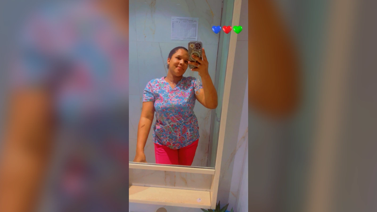 Woman takes a mirror selfie in a bathroom, wearing a blue patterned top and bright pink pants.