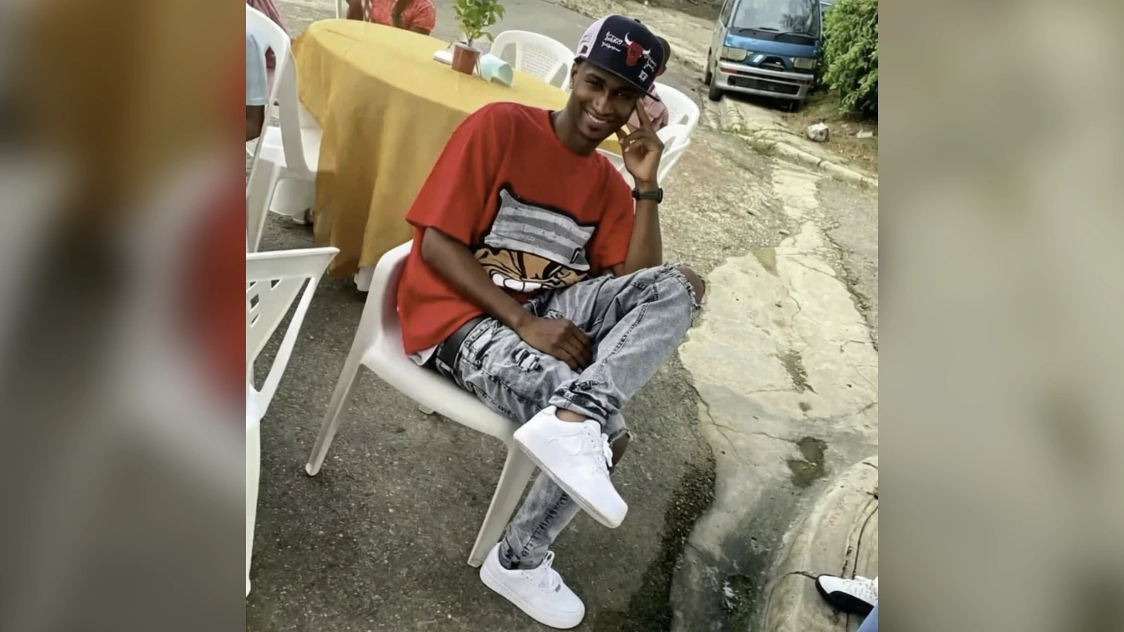 Young man sitting outdoors on a white plastic chair, wearing a red T‑shirt, ripped jeans, white sneakers, and a cap, smiling at the camera.