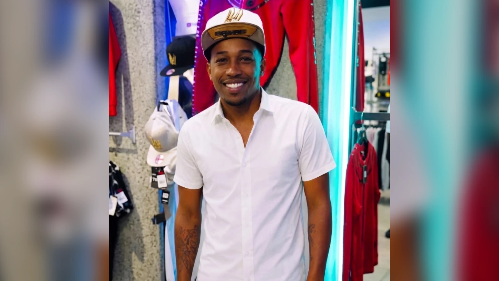 Smiling man in a white short-sleeve shirt and beige cap standing in a clothing store with hats and colorful jackets behind him.