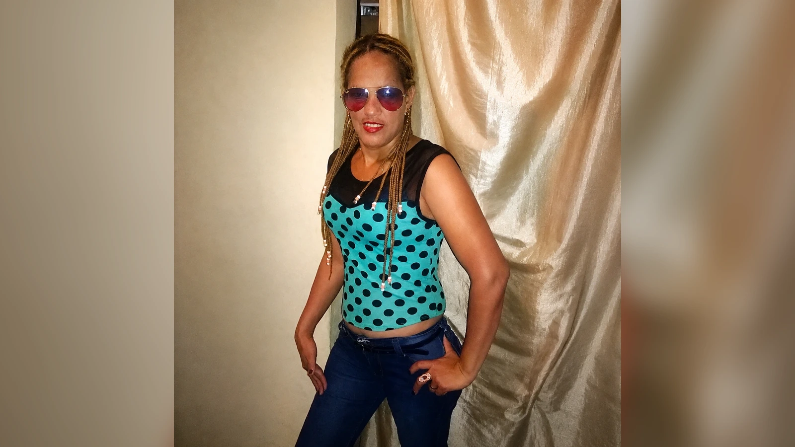 Portrait of a woman in a teal polka-dot sleeveless top and blue jeans, wearing aviator sunglasses and braided hair, posing with hands on hips.