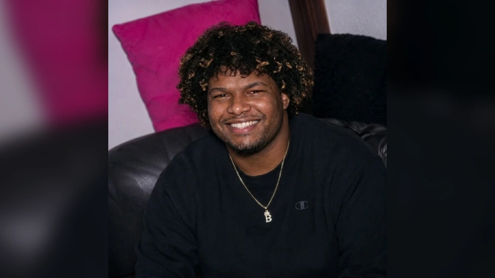 Smiling man with curly hair wearing a black shirt and gold necklace, seated on a leather couch with a pink pillow behind him.