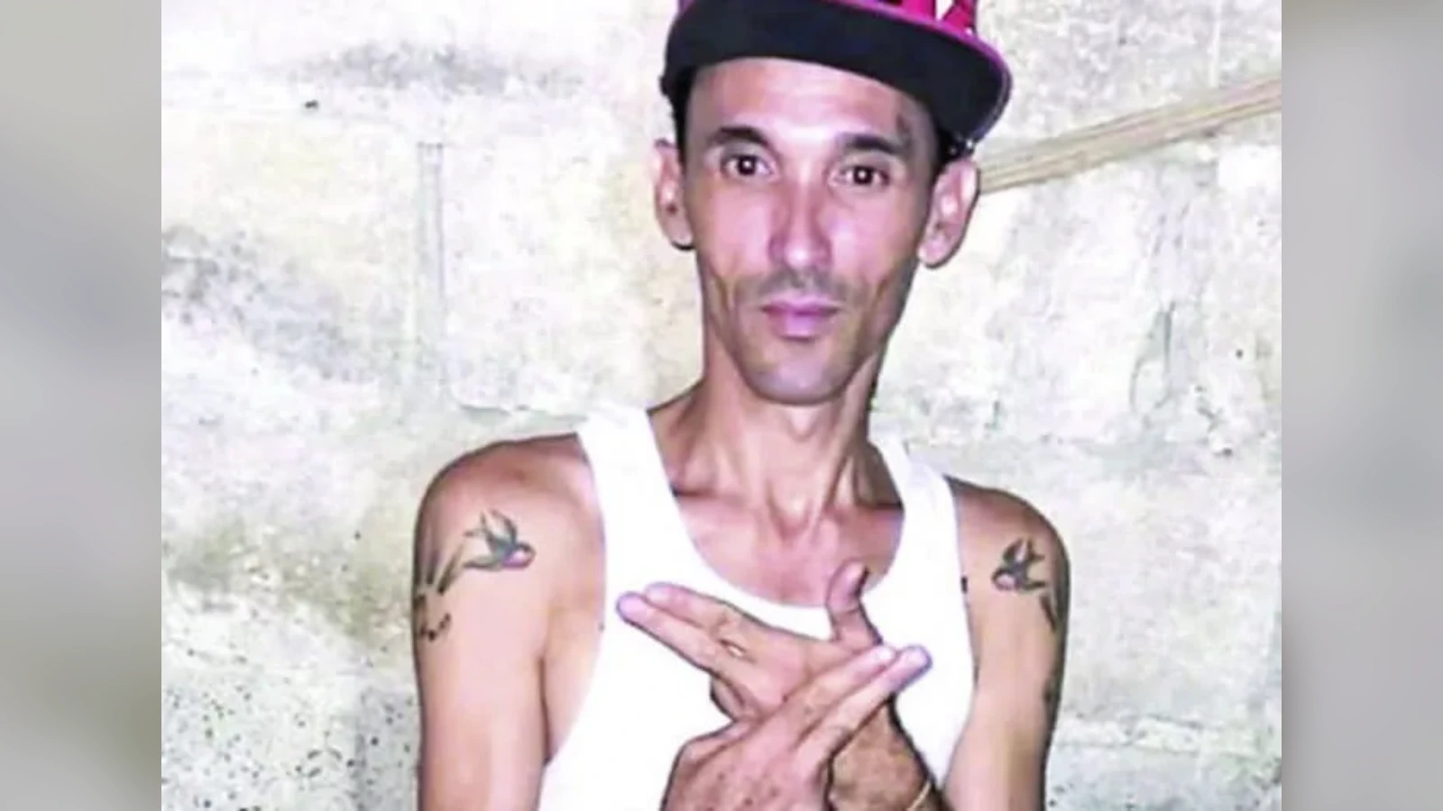 Young man wearing a white tank top and black cap with pink brim, standing against a concrete wall with tattooed arms crossed over his chest.