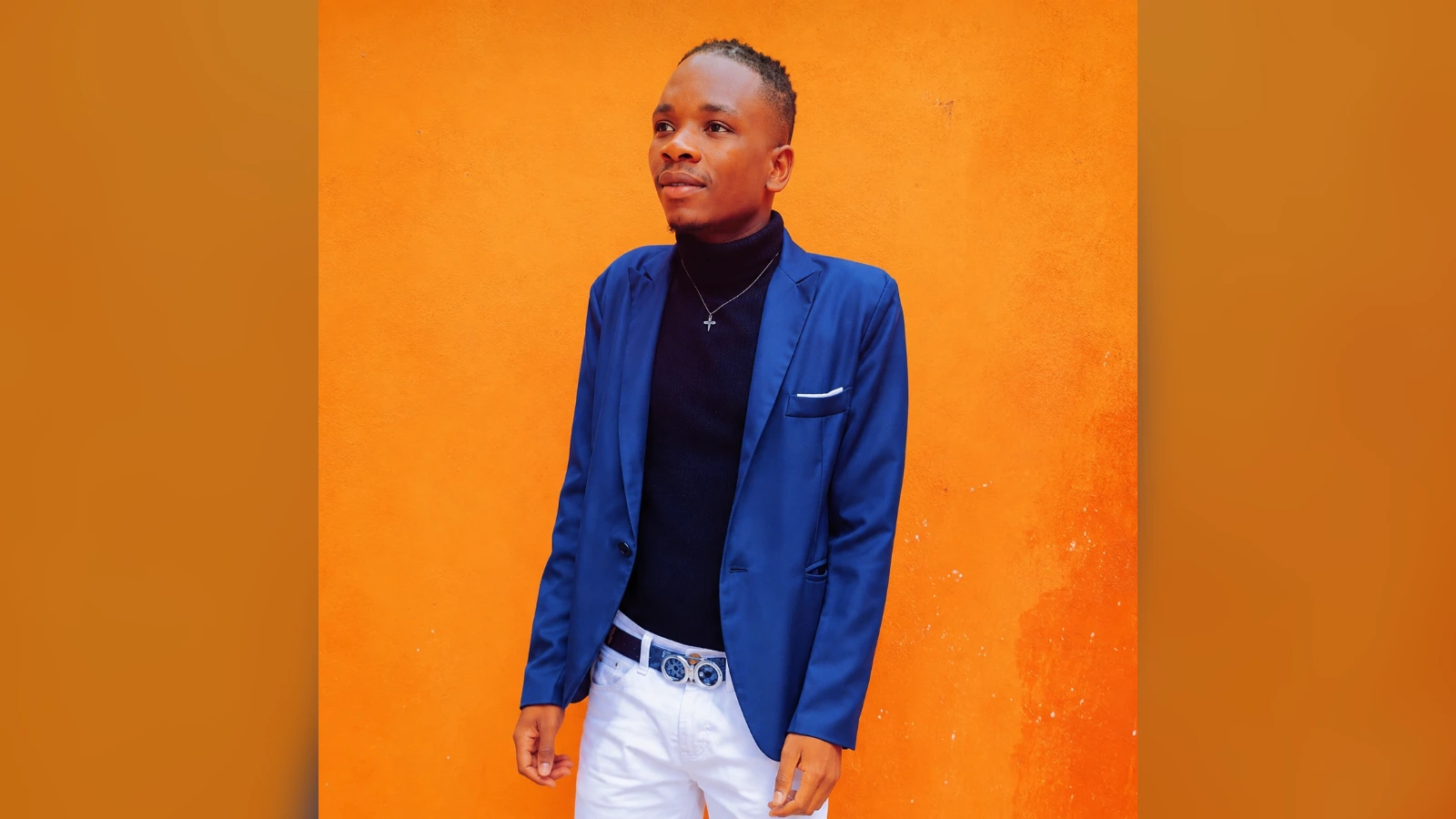 Young man in a blue blazer, black turtleneck, and white pants stands against a bright orange wall.