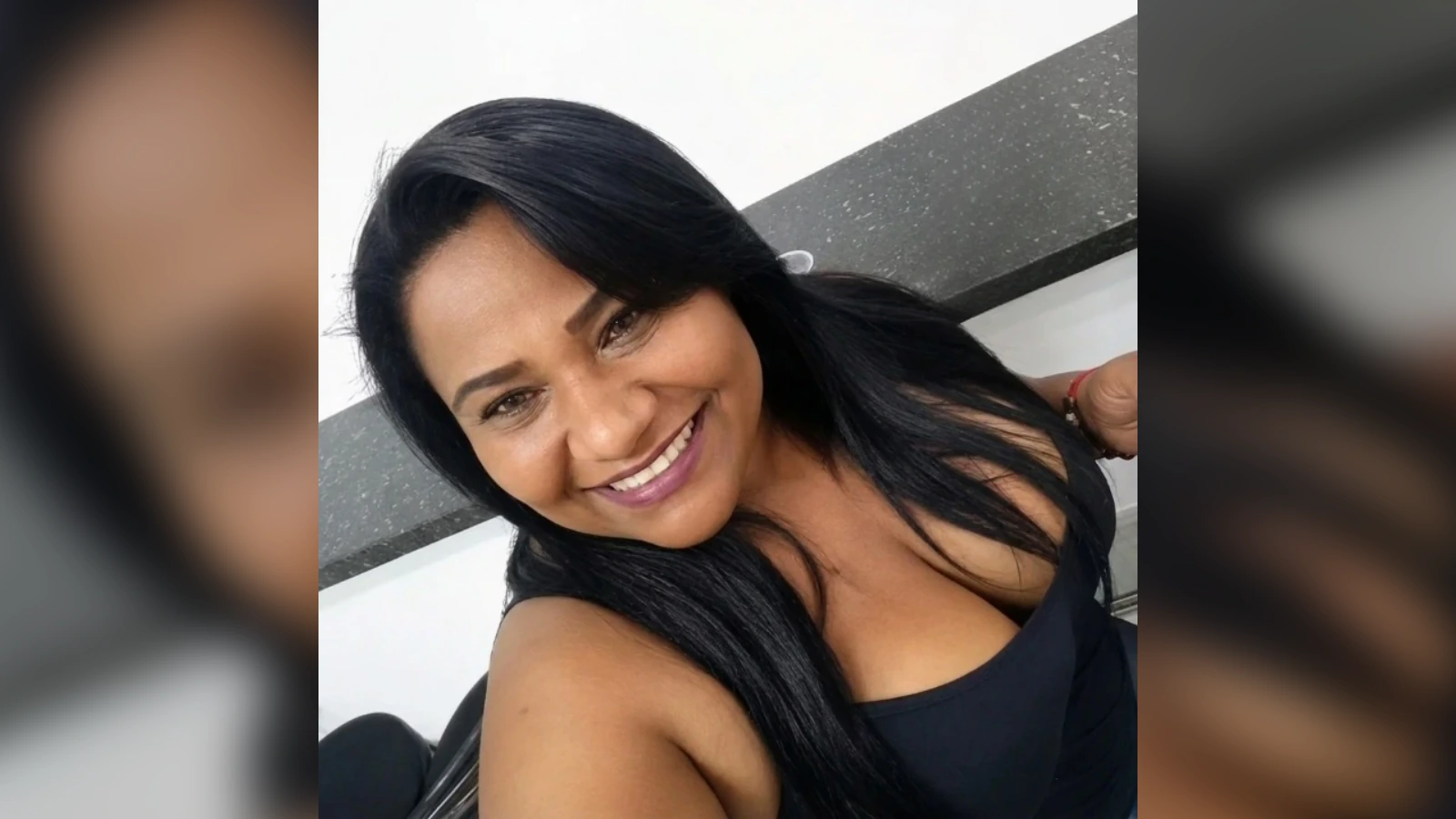 Smiling woman taking a selfie, long black hair, wearing a black sleeveless top.