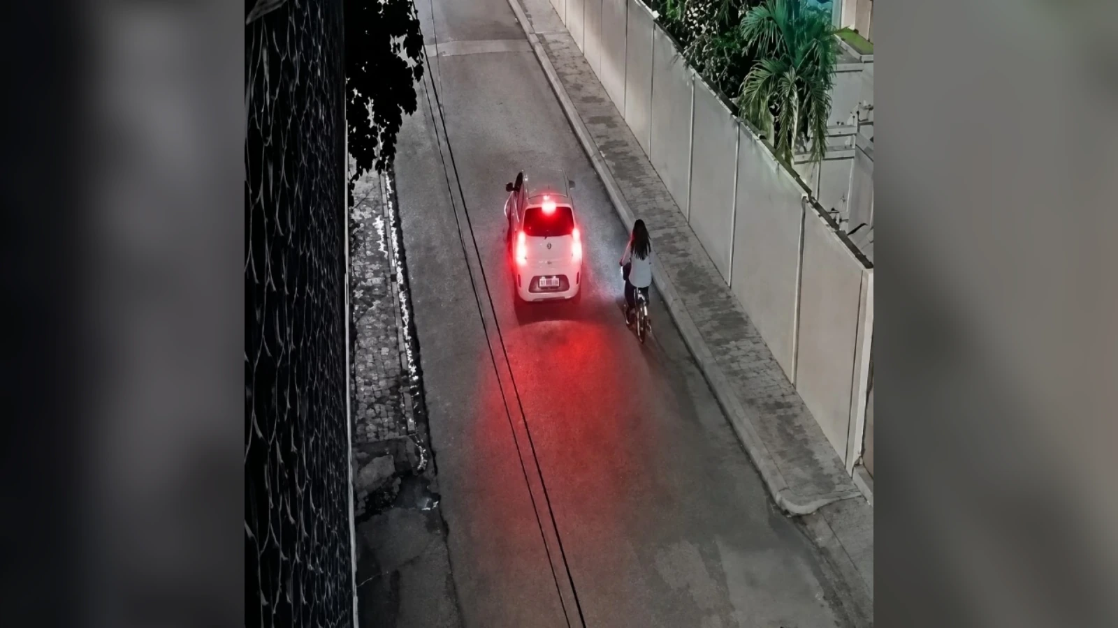 White hatchback stopped on a narrow street at night with a cyclist beside it; red brake lights reflect on the wet road behind them.