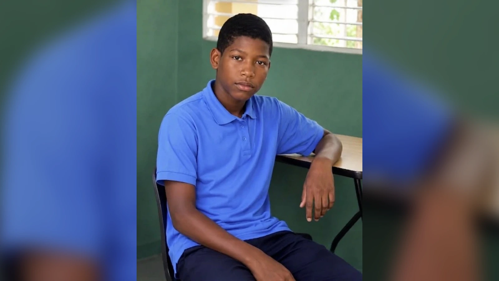 Teenage boy in a blue polo shirt sits at a desk in a classroom, looking at the camera.