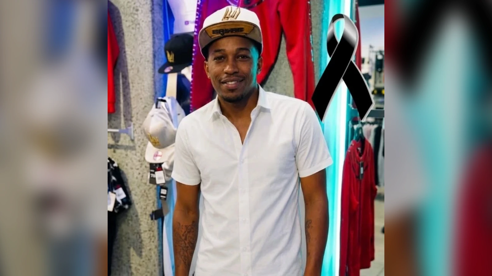 Smiling man in a white short-sleeve shirt and cap inside a clothing store, with hats and clothing on display in the background and a black ribbon visible.