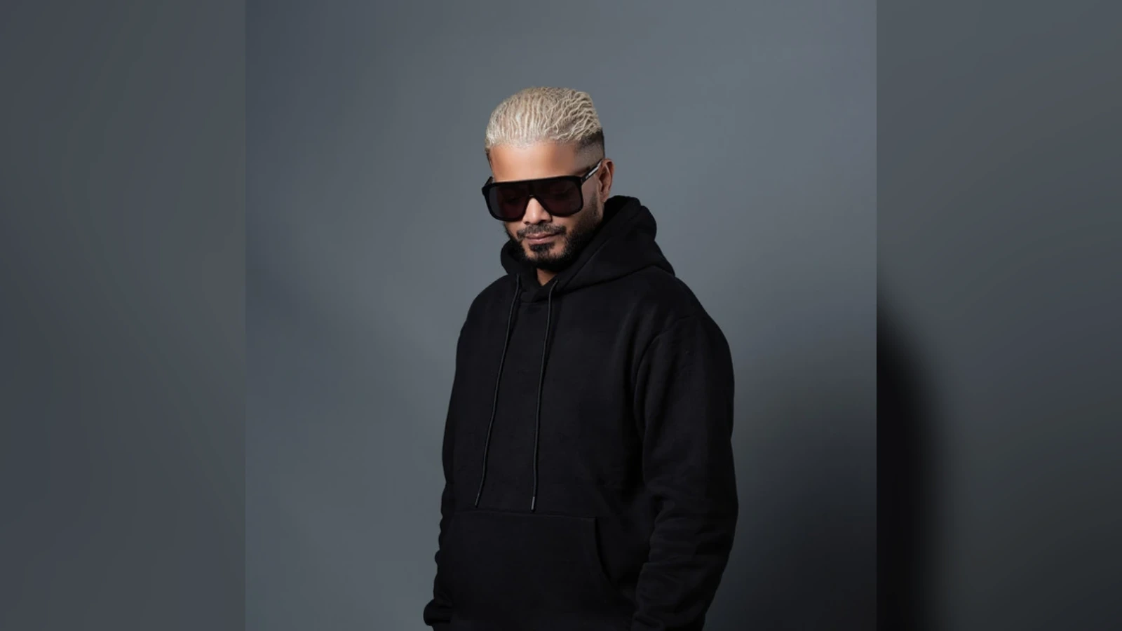 Man with bleached blond hair wearing a black hoodie and large dark sunglasses, posing against a gray studio background.