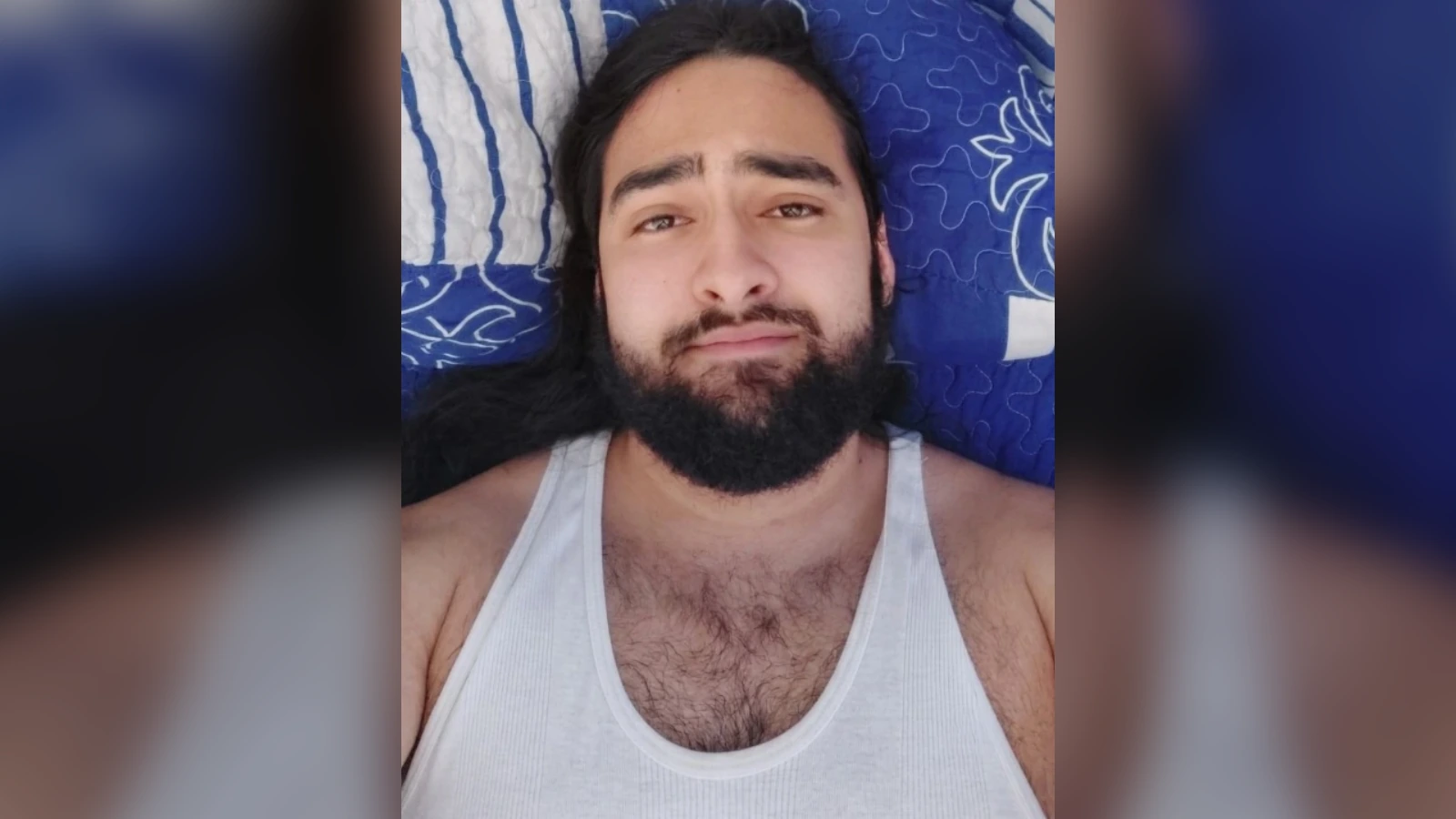 Bearded man wearing a white tank top, lying on a bed and looking at the camera.