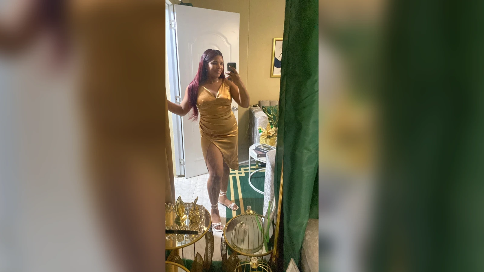 Woman in a gold satin dress posing for a mirror selfie in a living room with green curtains and gold decor, showing a thigh-high slit.