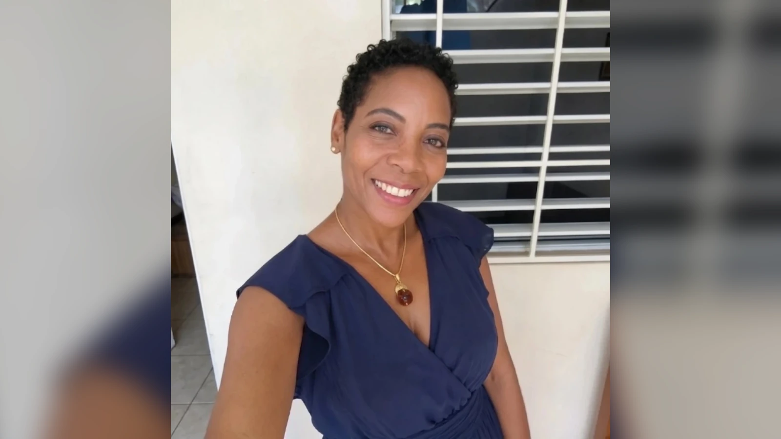 Smiling woman with short curly hair in a navy dress and gold pendant, standing beside a white wall and a barred window.