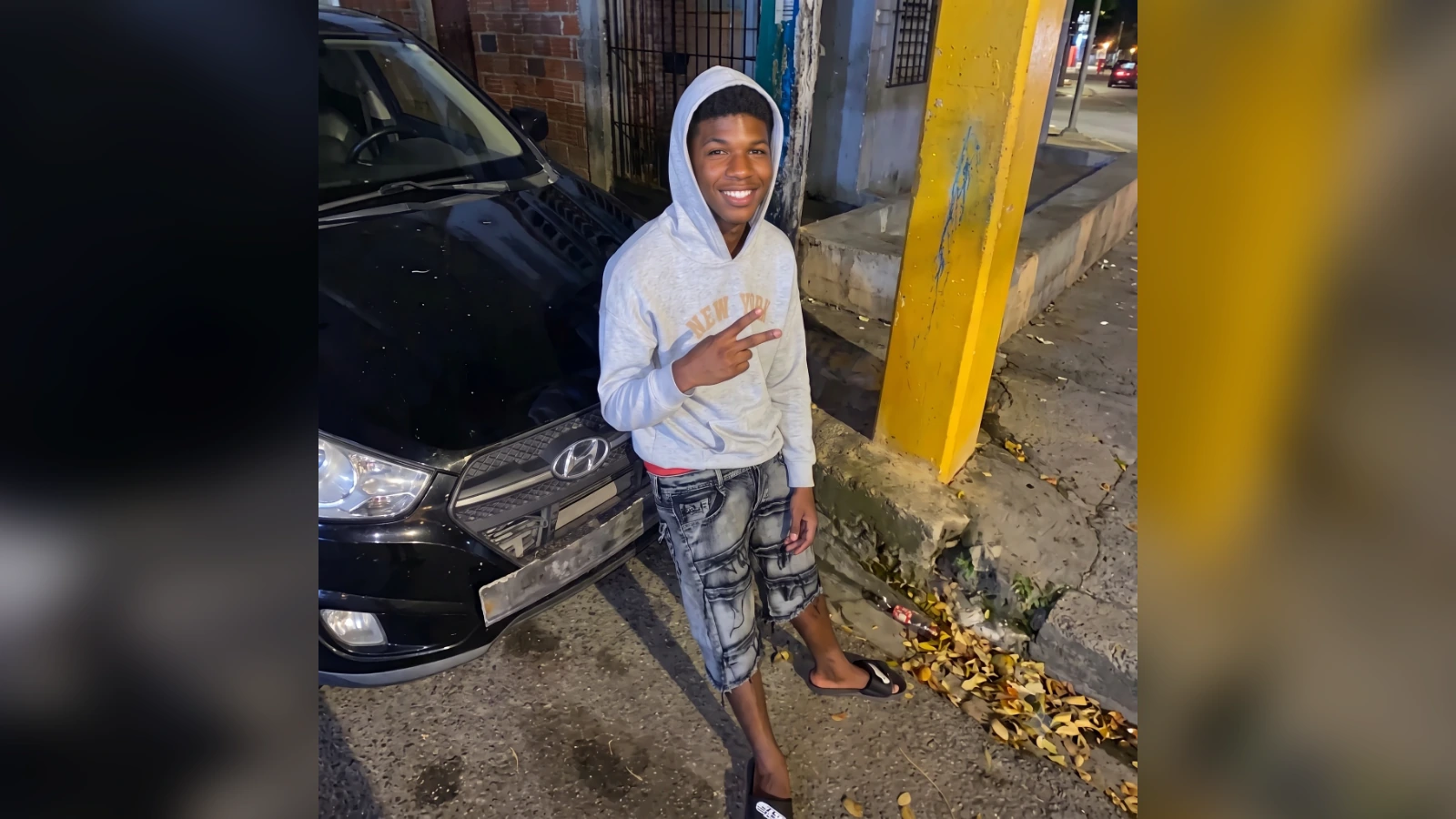 Young person in a light gray hoodie and shorts posing with a peace sign beside a parked black car at night on a city street.