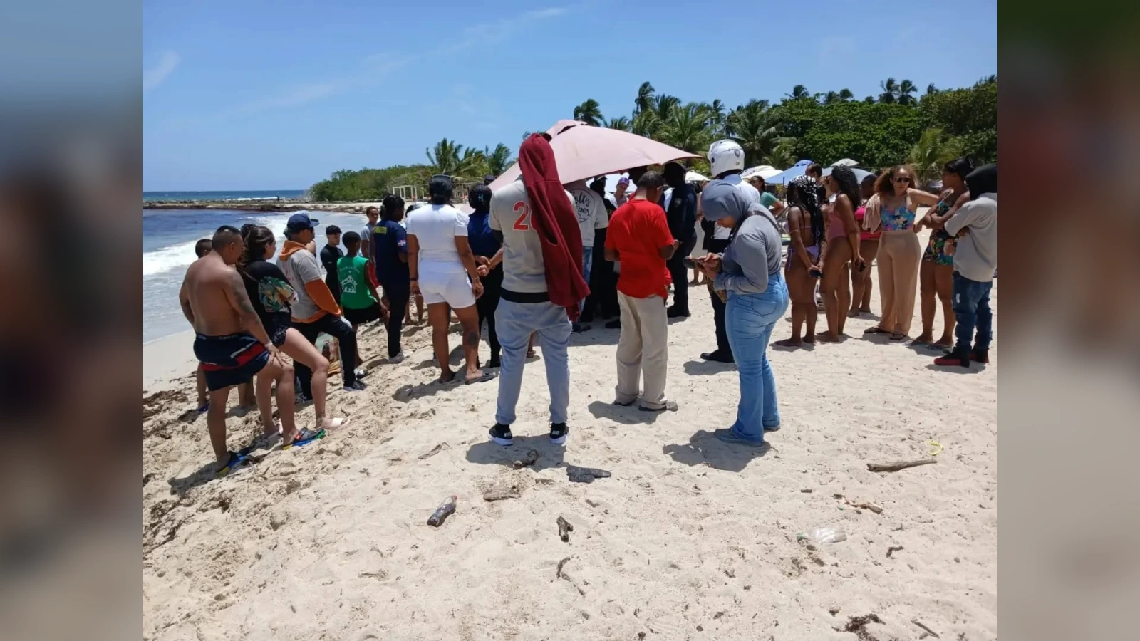 Crowd of beachgoers standing in a circle on the sandy shore under a pink umbrella, with palm trees and ocean in the background.